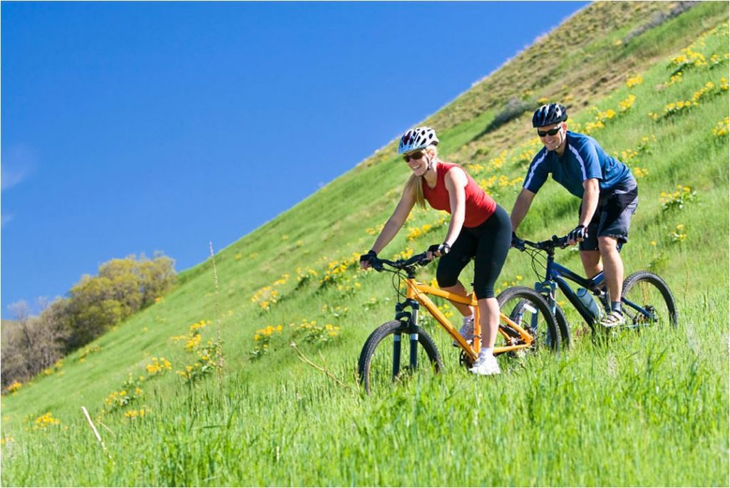 Aktivurlaub in Oberbayern: Zwei Radfahrer mit Helmen genießen eine Mountainbike-Tour über einen sonnigen, grünen Grashang vor strahlend blauem Himmel – Campingplatz Kaiser Camping in Bad Feilnbach.