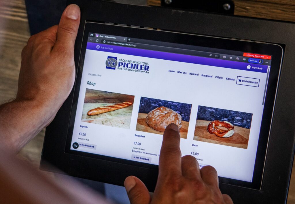 Eine Person nutzt ein Tablet, um Backwaren im Online-Shop der Bäckerei Pichler zu bestellen. Auf dem Bildschirm sind ein Baguette, ein Bauernbrot und eine Breze mit den jeweiligen Preisen zu sehen. Ein Finger tippt gerade auf das Bild des Bauernbrots.
