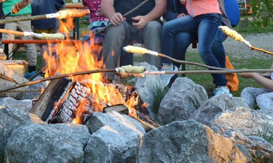 Eine Gruppe von Menschen sitzt gemütlich am Lagerfeuer und backt Stockbrot auf dem Campingplatz Kaiser Camping in Bad Feilnbach.