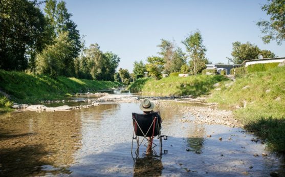 Idyllische Abkühlung im Sommer: Ein Camper mit Hut genießt die Ruhe in einem Klappstuhl mitten im seichten Flussbett des Jenbachs – Campingplatz Kaiser Camping in Bad Feilnbach.