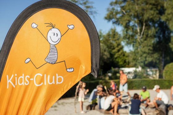 Kinderanimation beim Camping: Im Vordergrund ist das Logo des "Kids-Club" zu sehen, während im unscharfen Hintergrund eine Gruppe von Kindern und Betreuern zusammensitzt – Campingplatz Kaiser Camping in Bad Feilnbach.