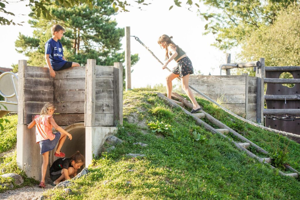 Abwechslungsreicher Kinderspielplatz im Kids-Club: Ein Mädchen klettert ein Seil am Grashügel hinauf, während andere Kinder am Holzpodest und beim Tunnel spielen – Campingplatz Kaiser Camping in Bad Feilnbach.