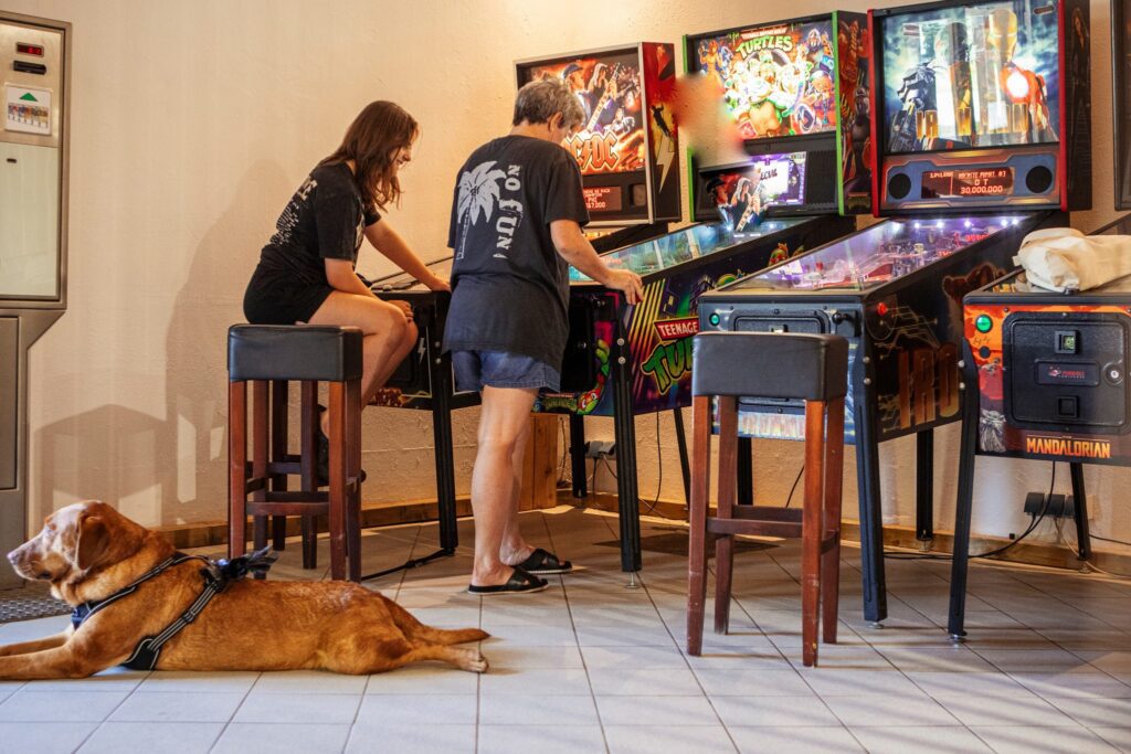 Freizeitspaß für Groß und Klein bei jedem Wetter: Eine Jugendliche und eine ältere Frau stehen an bunt leuchtenden Flipperautomaten in der Spielehalle auf dem Campingplatz Kaiser Camping in Bad Feilnbach.