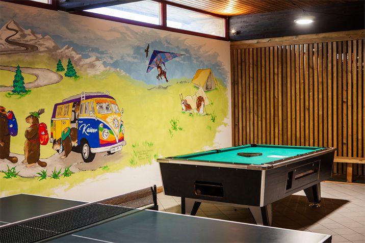 Indoor-Aktivitäten für Regentage: Ein grüner Billardtisch steht neben einer Tischtennisplatte vor einer großen, fröhlichen Wandmalerei mit Camping-Motiven – Campingplatz Kaiser Camping in Bad Feilnbach.