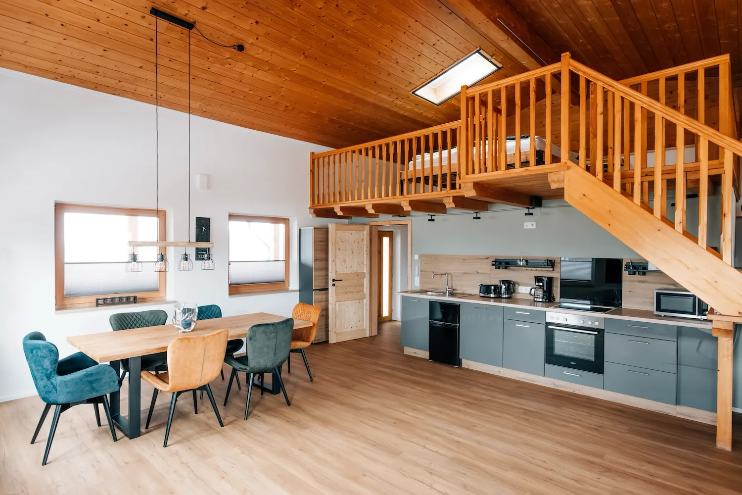 Wohn-Ess-Küchen-Bereich der Ferienwohnung Kaiser Franz Josef: moderner Wohnbereich mit blauem Sessel, grauer Einbauküche und Holzstiege zur Galerie - Kaiser Campingplatz in Bad Feilnbach