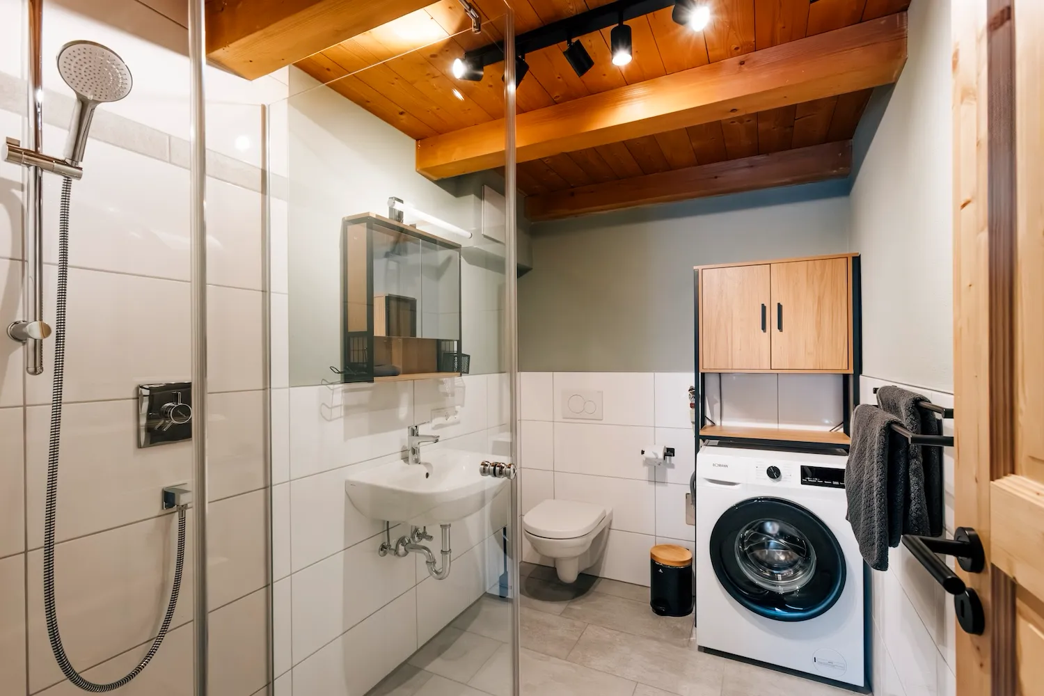 Modernes Badezimmer der Ferienwohnung Kaiser Franz Josef mit Dusche, Waschmaschine, WC und hellen Fliesen unter Holzdecke - Kaiser Campingplatz in Bad Feilnbach