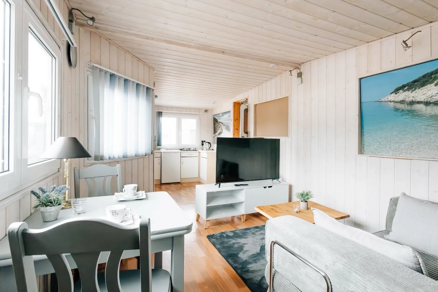 Wohn- und Essbereich im festen Anbau der Camping-Suite Karl Albrecht: Sofa, Esstisch und Flachbild-TV in hellem Raum mit weißer Holzverkleidung - Kaiser Campingplatz in Bad Feilnbach