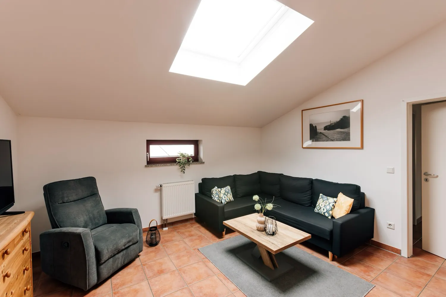 Wohnzimmer der Ferienwohnung König Ludwig mit dunklem Sofa, Sessel und Dachfenster – gemütliche Atmosphäre unter der Dachschräge - Kaiser Campingplatz in Bad Feilnbach