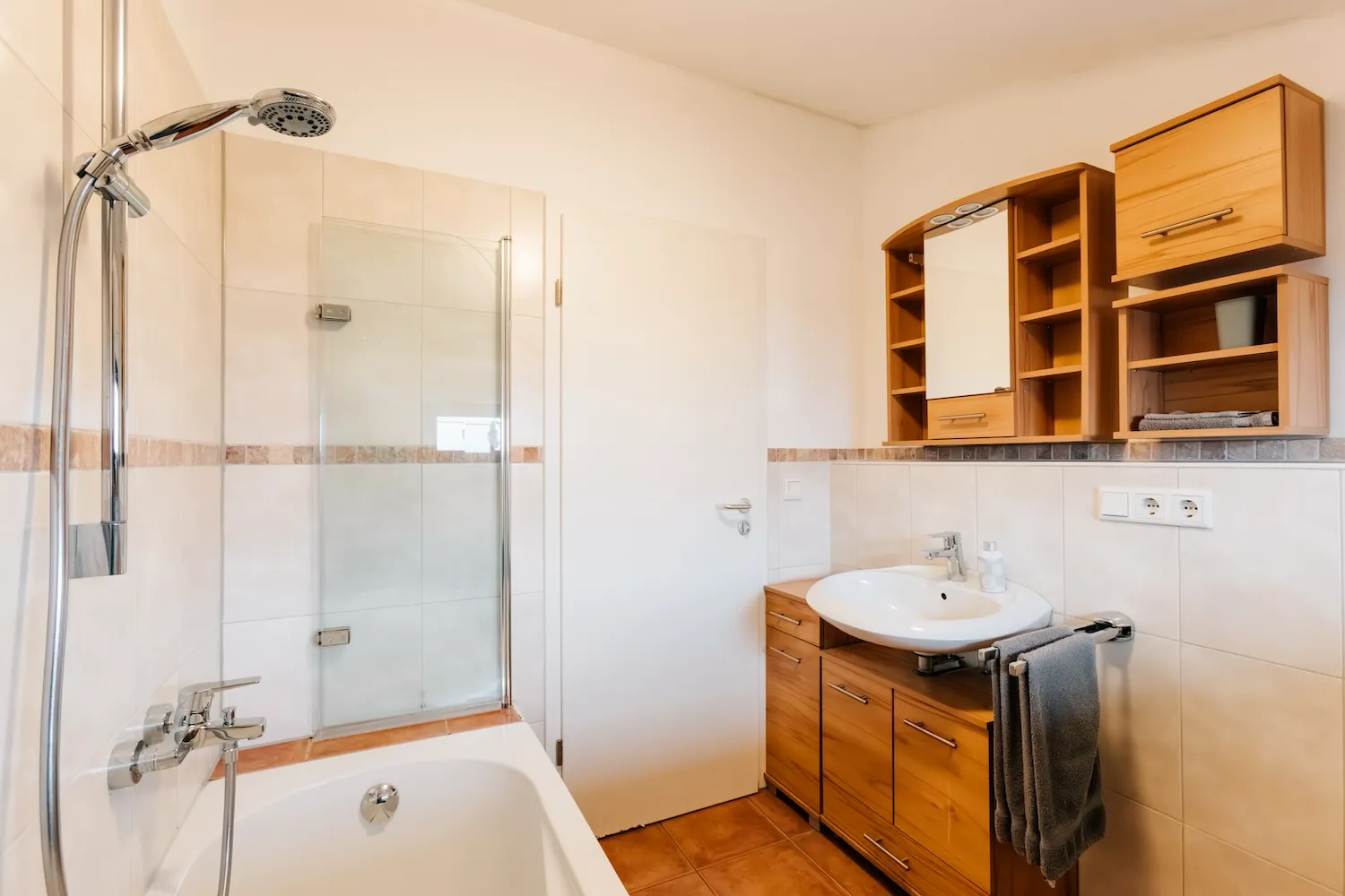 Badezimmer der Ferienwohnung König Ludwig: Badewanne mit Duschkombination, Holzregal und helle Fliesen – ruhige, klassische Ausstattung - Kaiser Campingplatz in Bad Feilnbach
