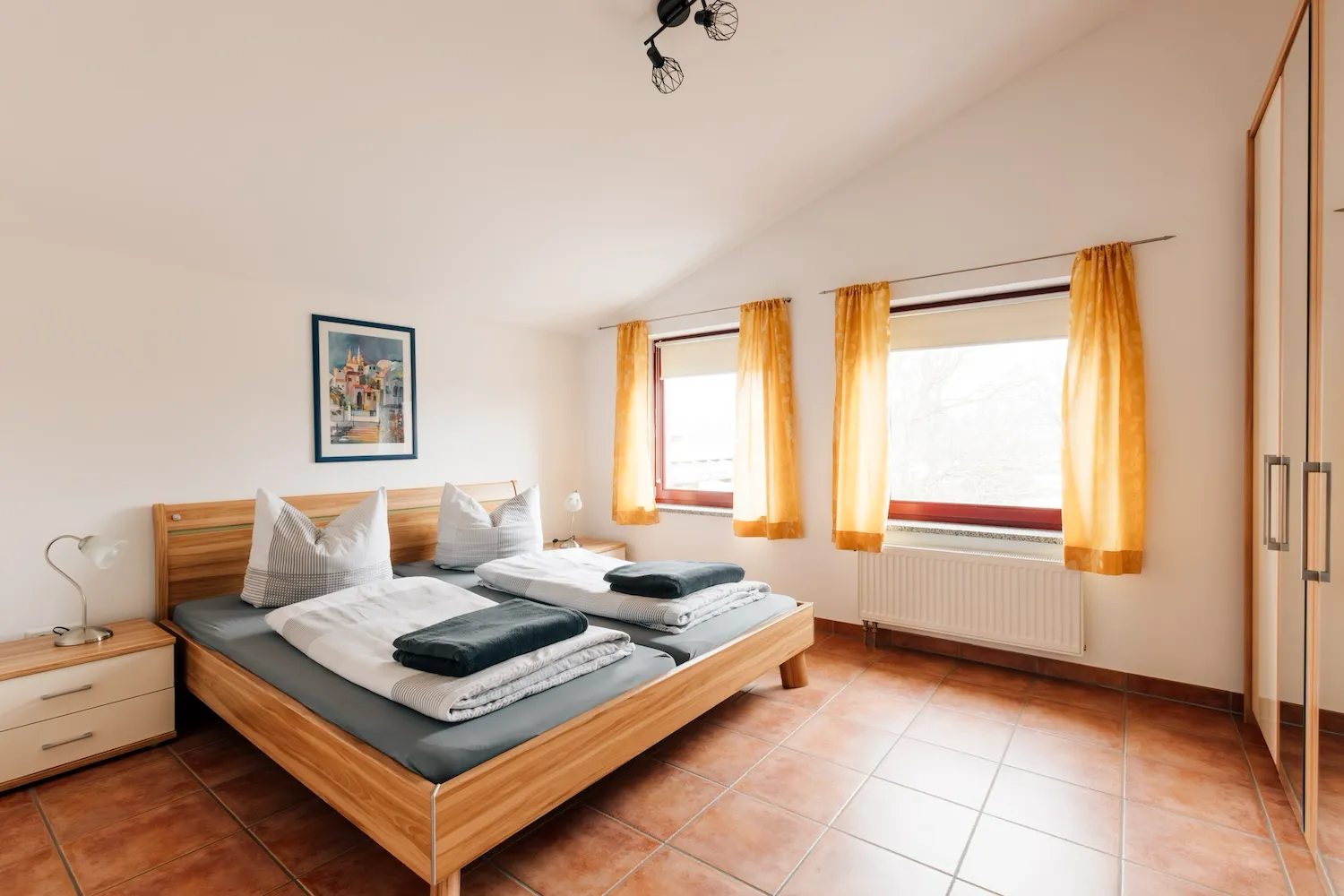 Schlafzimmer der Ferienwohnung König Ludwig mit Doppelbett in Holzrahmen, Terrakottaboden und warmem Licht durch gelbe Vorhänge - Kaiser Campingplatz in Bad Feilnbach