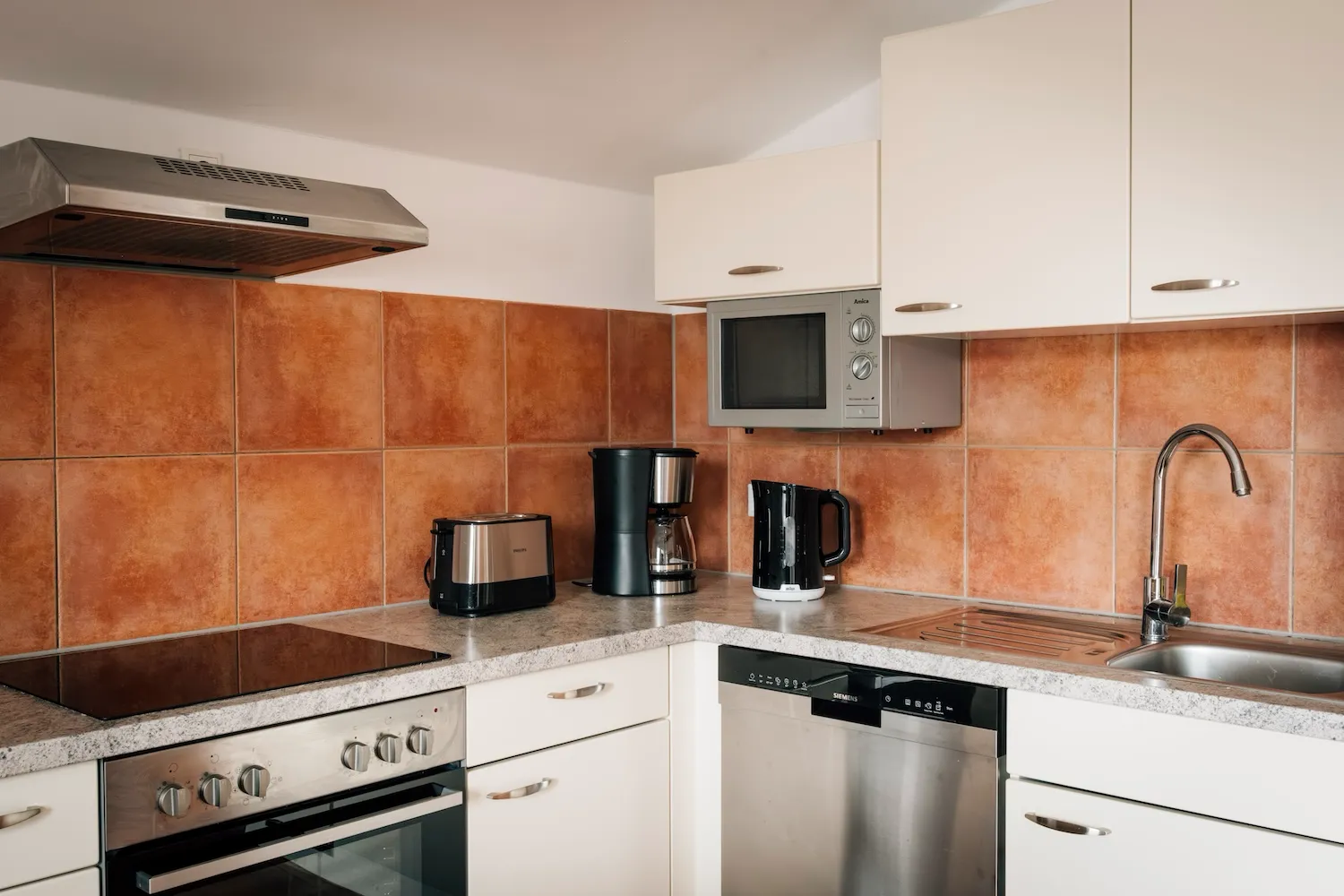 Küchendetail der Ferienwohnung König Ludwig: Cerankochfeld, Mikrowelle, Kaffeemaschine und Geschirrspüler auf klassischem Terrakottaboden - Kaiser Campingplatz in Bad Feilnbach
