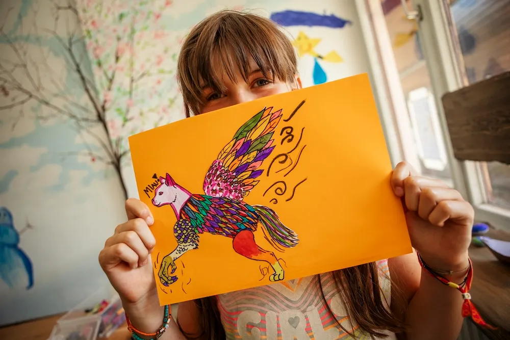 Basteln und kreativ sein im Urlaub: Ein Kind zeigt ein selbst gestaltetes, farbenfrohes Bild auf orangefarbenem Papier im Innenbereich des Kids-Clubs auf dem Campingplatz Kaiser Camping in Bad Feilnbach.