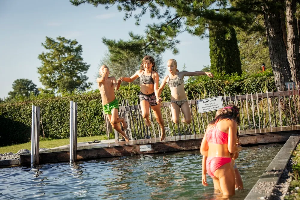 Spaß beim Baden im Sommer: Ein Junge und zwei Mädchen springen Hand in Hand ins klare Wasser des biologischen Schwimmteichs – Campingplatz Kaiser Camping in Bad Feilnbach.