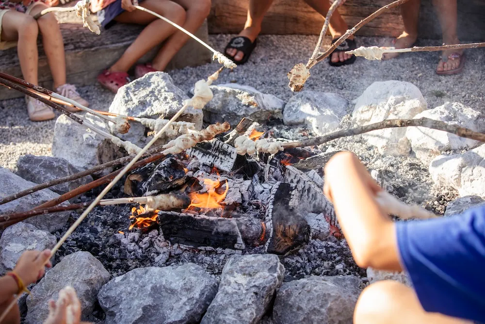Nahaufnahme einer gemauerten Feuerstelle, über der mehrere Kinder gemeinsam Stockbrot über der heißen Glut und den Flammen backen. Campingplatz Kaiser Camping in Bad Feilnbach „kids-club“