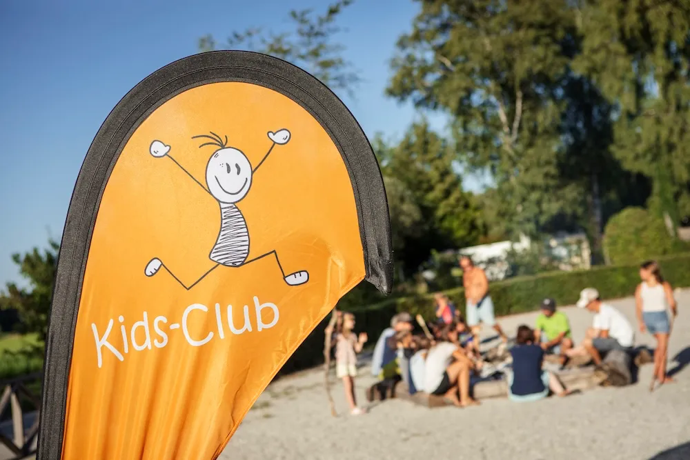 Blick auf die orangefarbene Strandfahne des „Kids-Club“ mit einem fröhlichen Strichmännchen-Logo. Im unscharfen Hintergrund versammelt sich eine Gruppe zur Kinderbetreuung rund um einen gemütlichen Lagerfeuerplatz. Campingplatz Kaiser Camping in Bad Feilnbach „kids-club“
