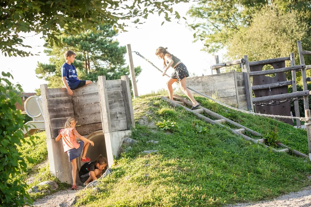 Eine lebhafte Szene auf dem Spielplatz: Jemand zieht sich an einem Seil den grünen Hügel hinauf, während andere beim Tunnelbauwerk klettern und den Spielbereich erkunden. Campingplatz Kaiser Camping in Bad Feilnbach Kids-Club