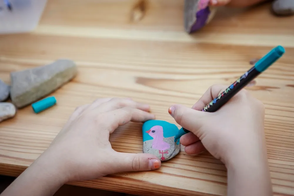Kreative Beschäftigung bei der Kinderbetreuung: Ein Kind verschönert einen Stein mit einem rosa Vogel auf blauem Hintergrund mithilfe eines Acrylmarkers auf einem Holztisch. Campingplatz Kaiser Camping in Bad Feilnbach Kids-Club