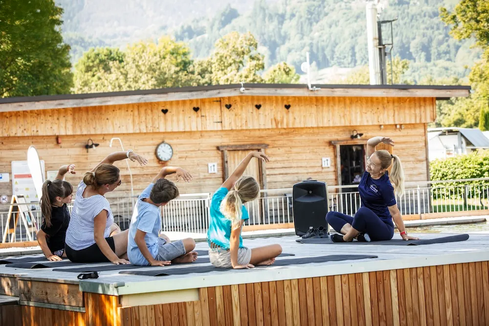 Fitness- und Gymnastikkurs beim Camping: Eine Gruppe macht gemeinsam Yoga- oder Dehnübungen auf Sportmatten vor einem Holzgebäude, angeleitet von einer Trainerin auf dem Campingplatz Kaiser Camping in Bad Feilnbach.